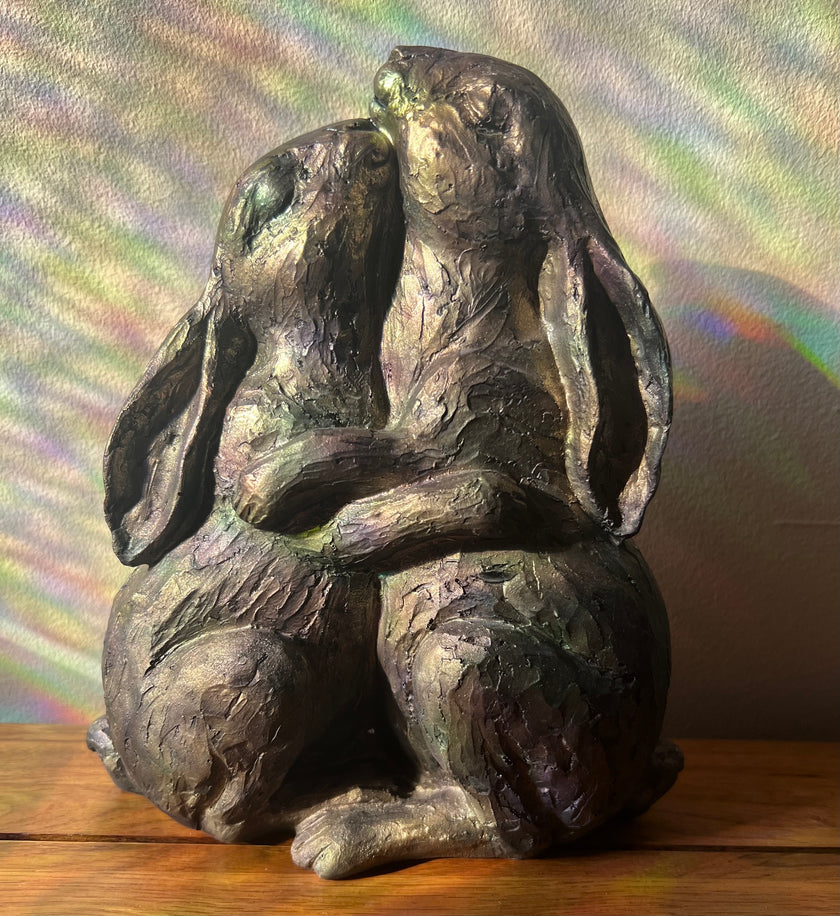 Bronze sculpture of two rabbits sitting together on a wooden surface with a rainbow gradient background.