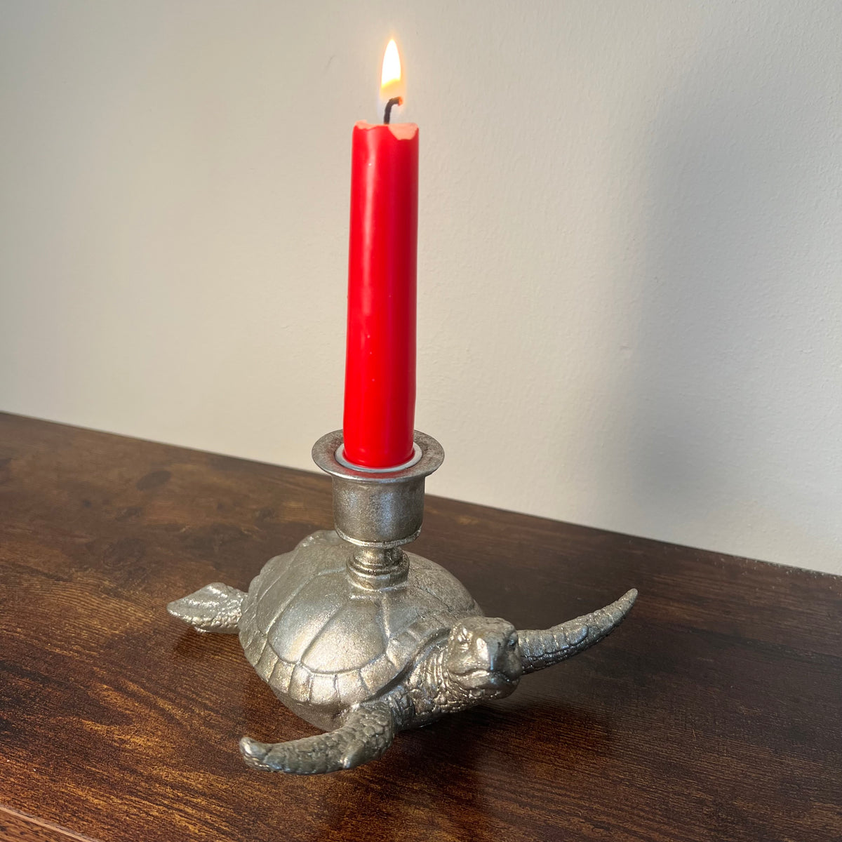 A rustic silver sea turtle candle holder, with a red taper candle, displayed on a dark oak shelf.